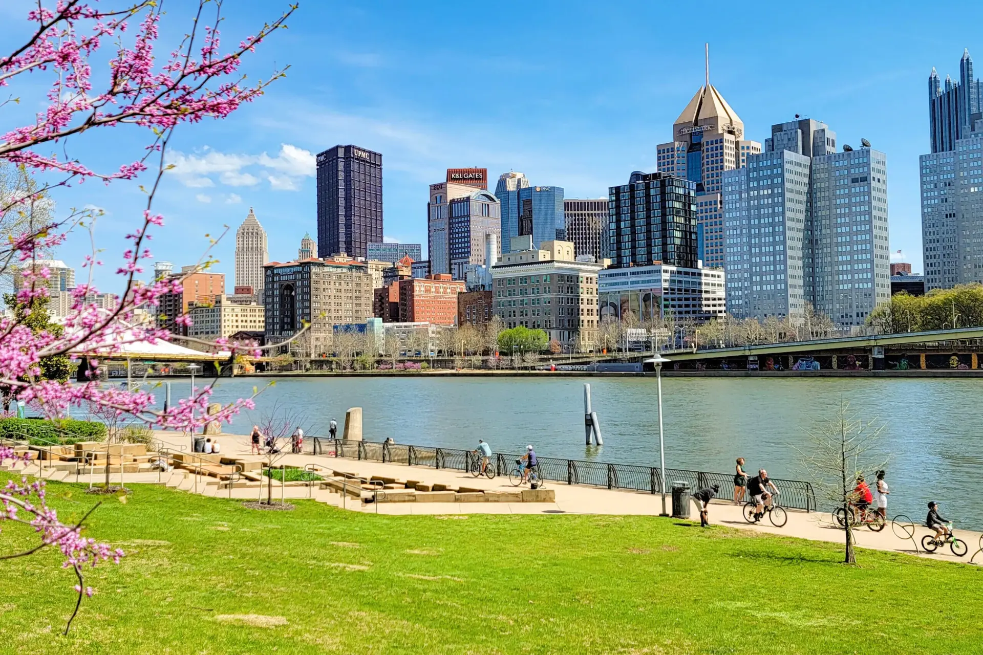 Cyclists on the riverfront trail across from the Downtown Pittsburgh skyline in spring blossom